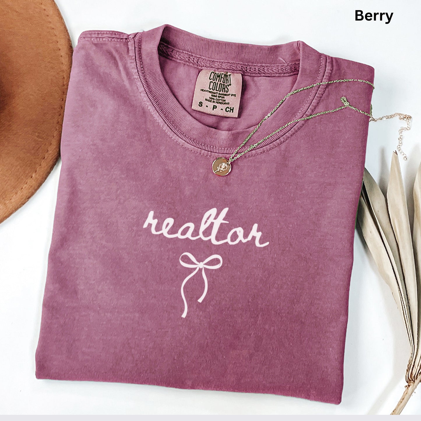 A pink t-shirt with the word "realtor" printed on it, along with a necklace and a brown hat, all placed on a white surface.