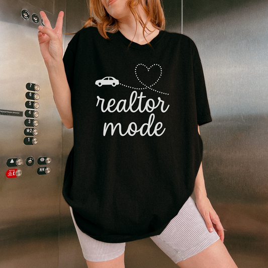 A person wearing a black t-shirt with the text "realtor mode" and a heart symbol, standing in front of an elevator.