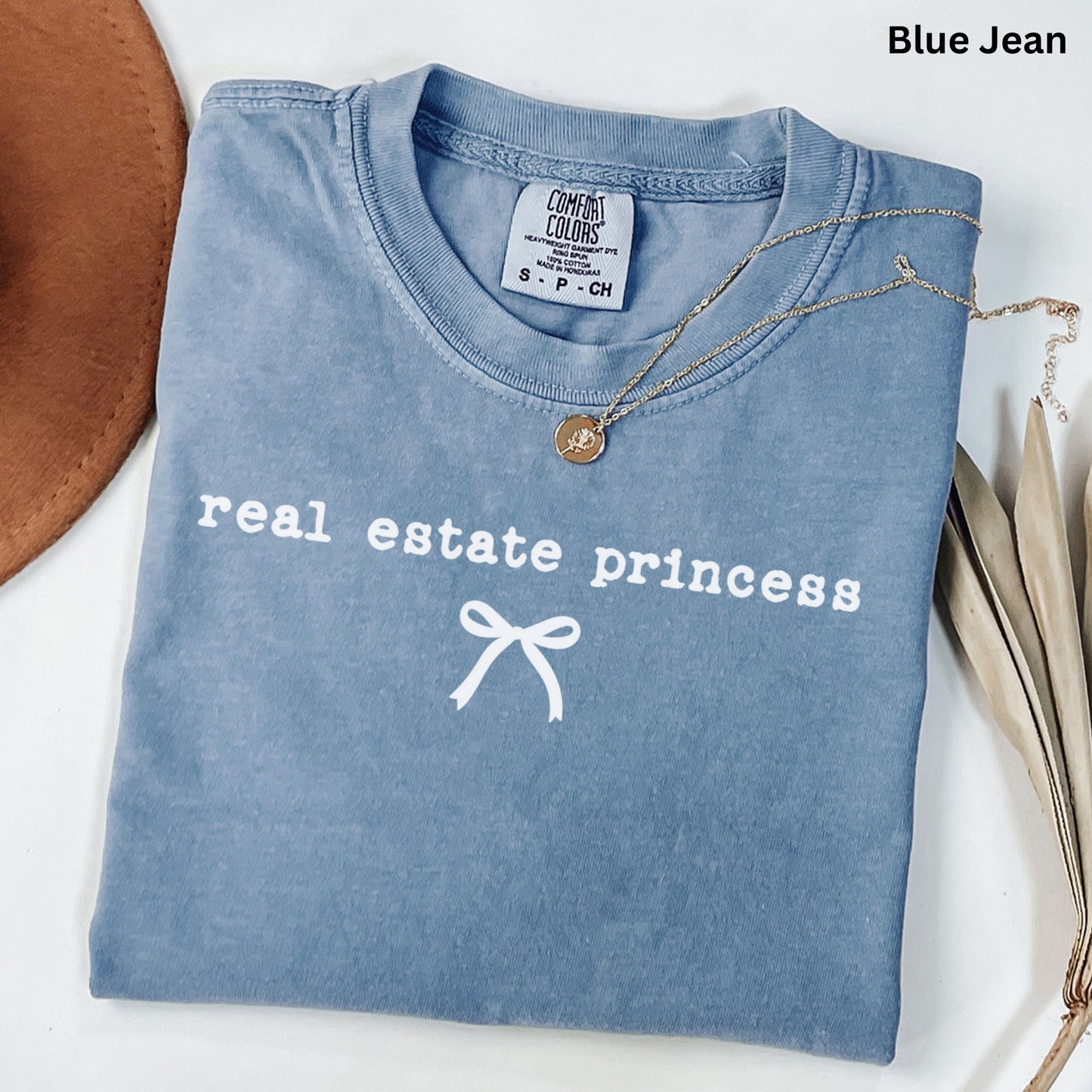 A blue t-shirt with the text "real estate princess" printed on it, along with a ribbon design. The t-shirt is laid out on a white surface, and there is a necklace and some leaves nearby.
