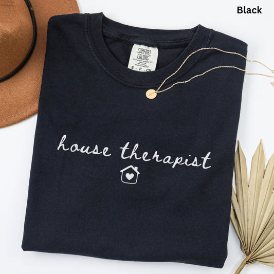 a black t-shirt with the text "house therapist" printed on it, along with a small house icon. The t-shirt is laid out on a white surface, and there is a brown hat and a leaf visible in the background.