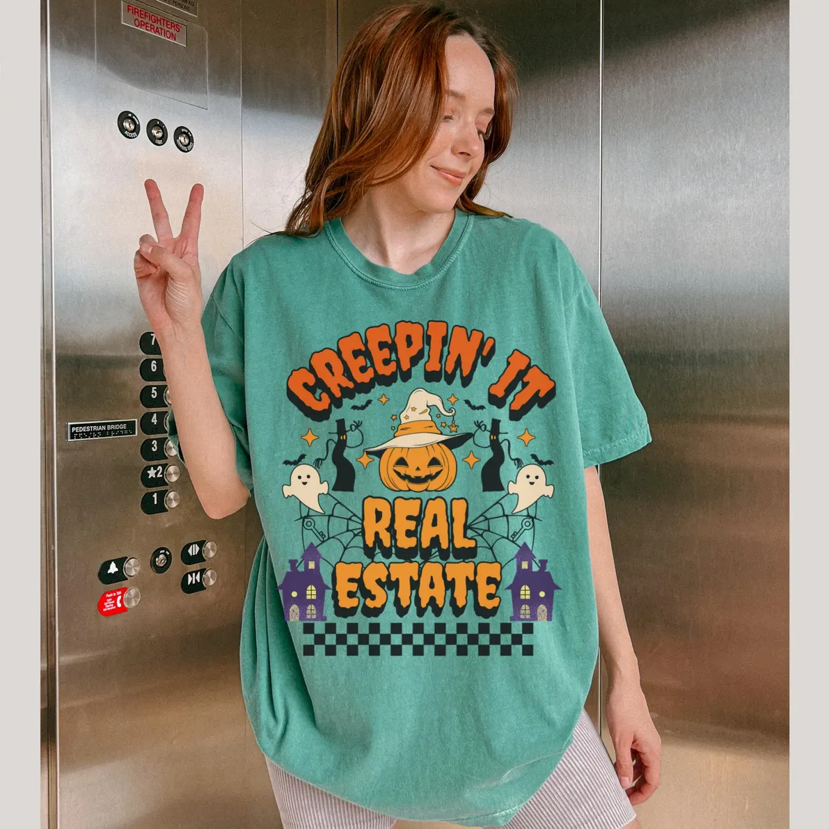 A woman wearing a green t-shirt with a Halloween-themed graphic and text, smiling and making a peace sign gesture.
