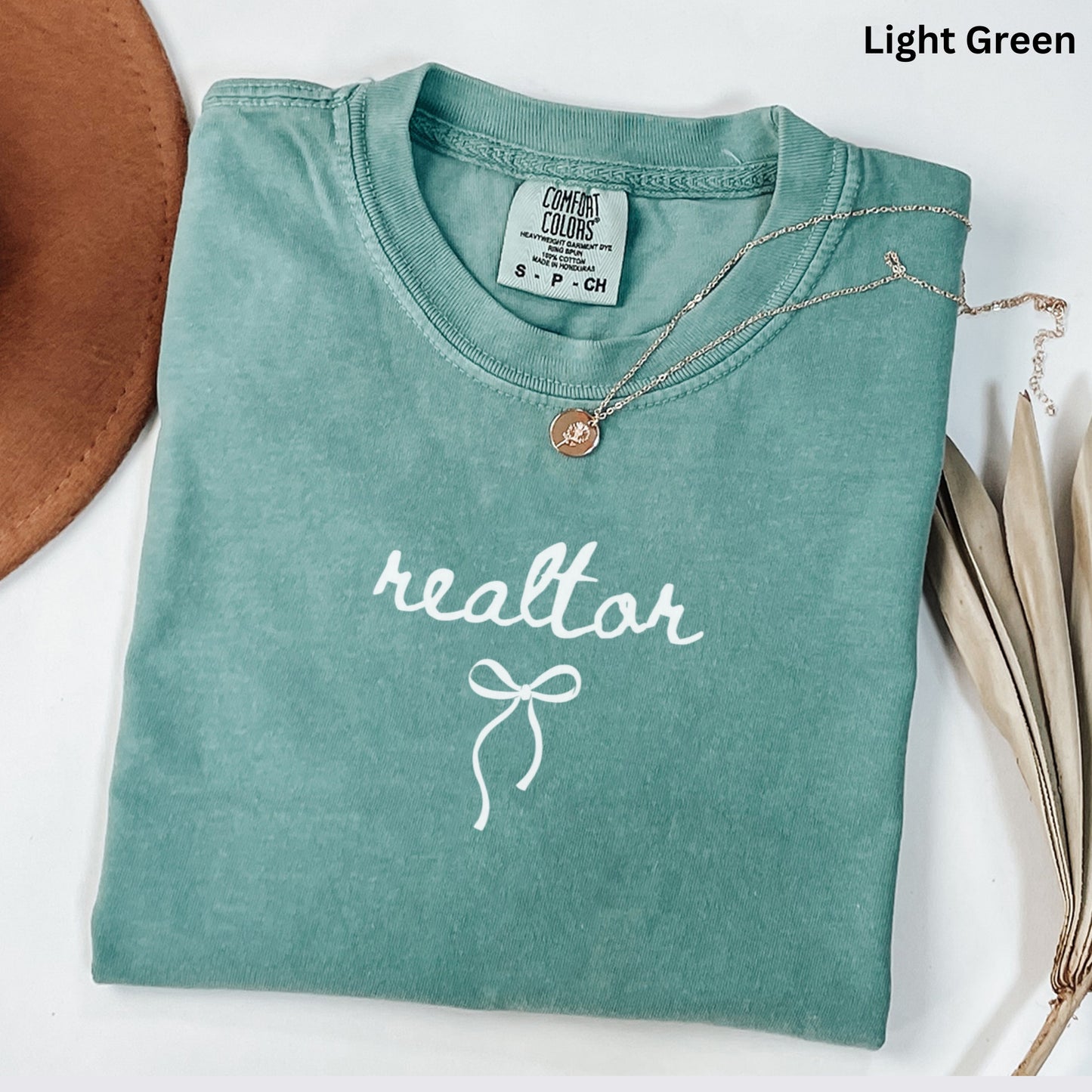 A light green t-shirt with the word "realtor" printed on it, along with a small gold pendant necklace. The t-shirt is folded and placed on a white surface, with a brown hat and some dried leaves visible in the background.