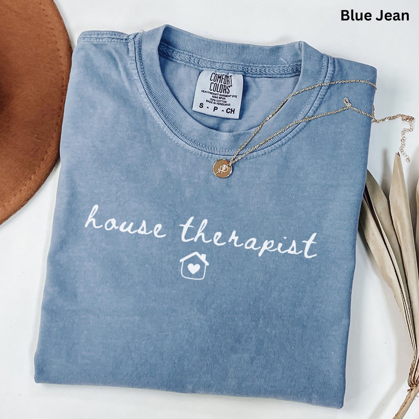 A blue t-shirt with the text "house therapist" printed on it, along with a small house icon. The t-shirt is folded and placed on a white surface, with a brown hat and some leaves visible in the background.