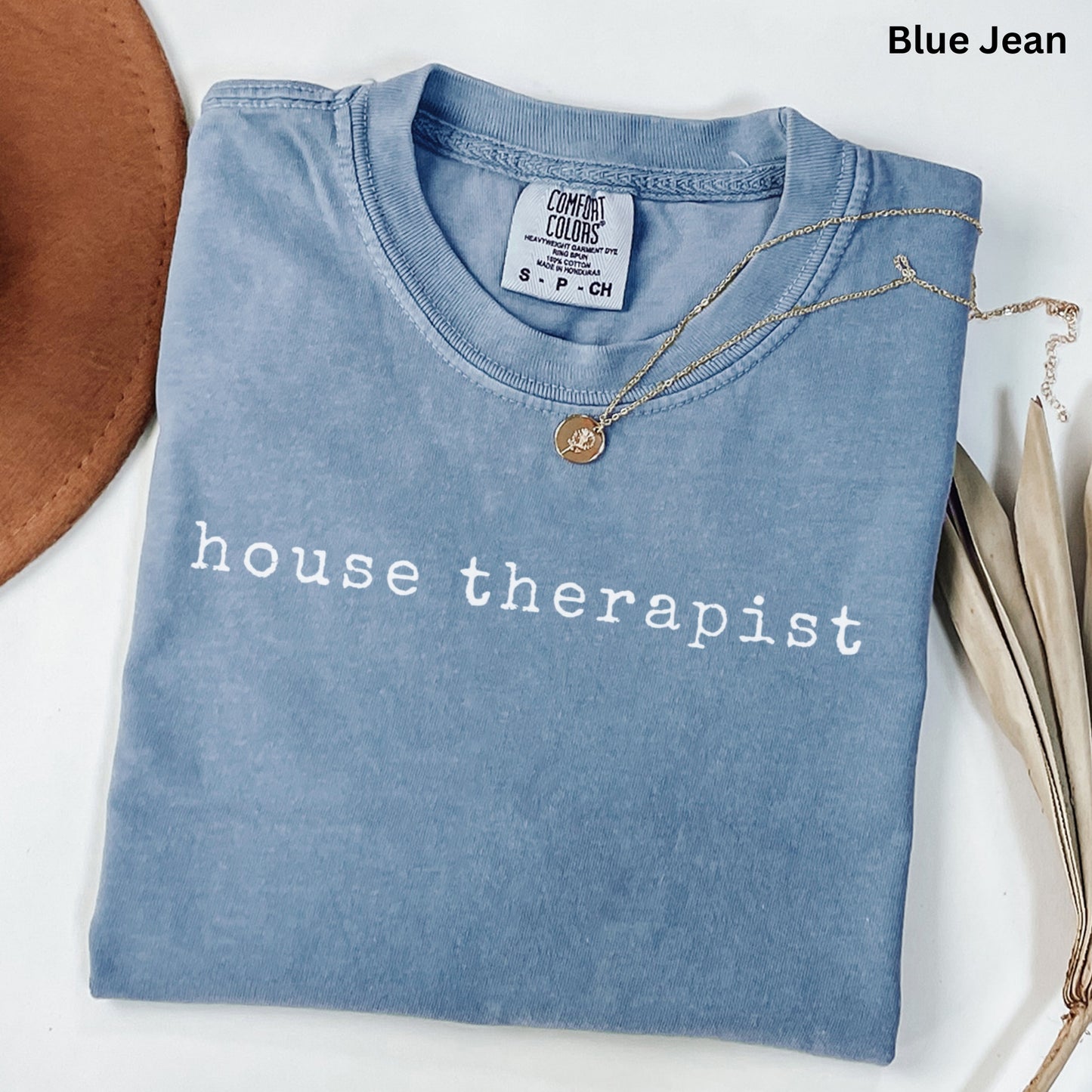 A blue t-shirt with the text "house therapist" printed on it, along with a necklace and some leaves.
