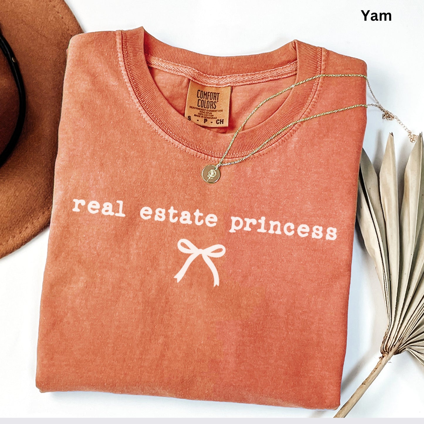 a red t-shirt with the text "real estate princess" printed on it, along with a bow design. The t-shirt is laid out on a white surface, and there is a brown hat and a plant in the background.