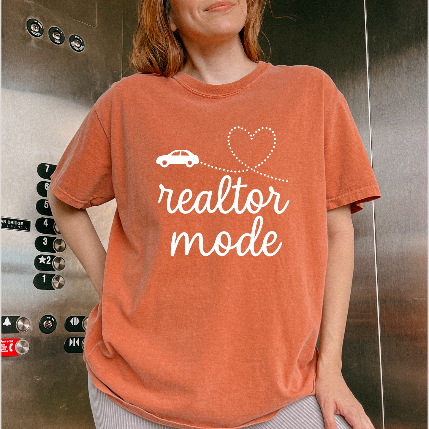 A woman wearing a red t-shirt with the text "realtor mode" printed on it, standing in front of an elevator.