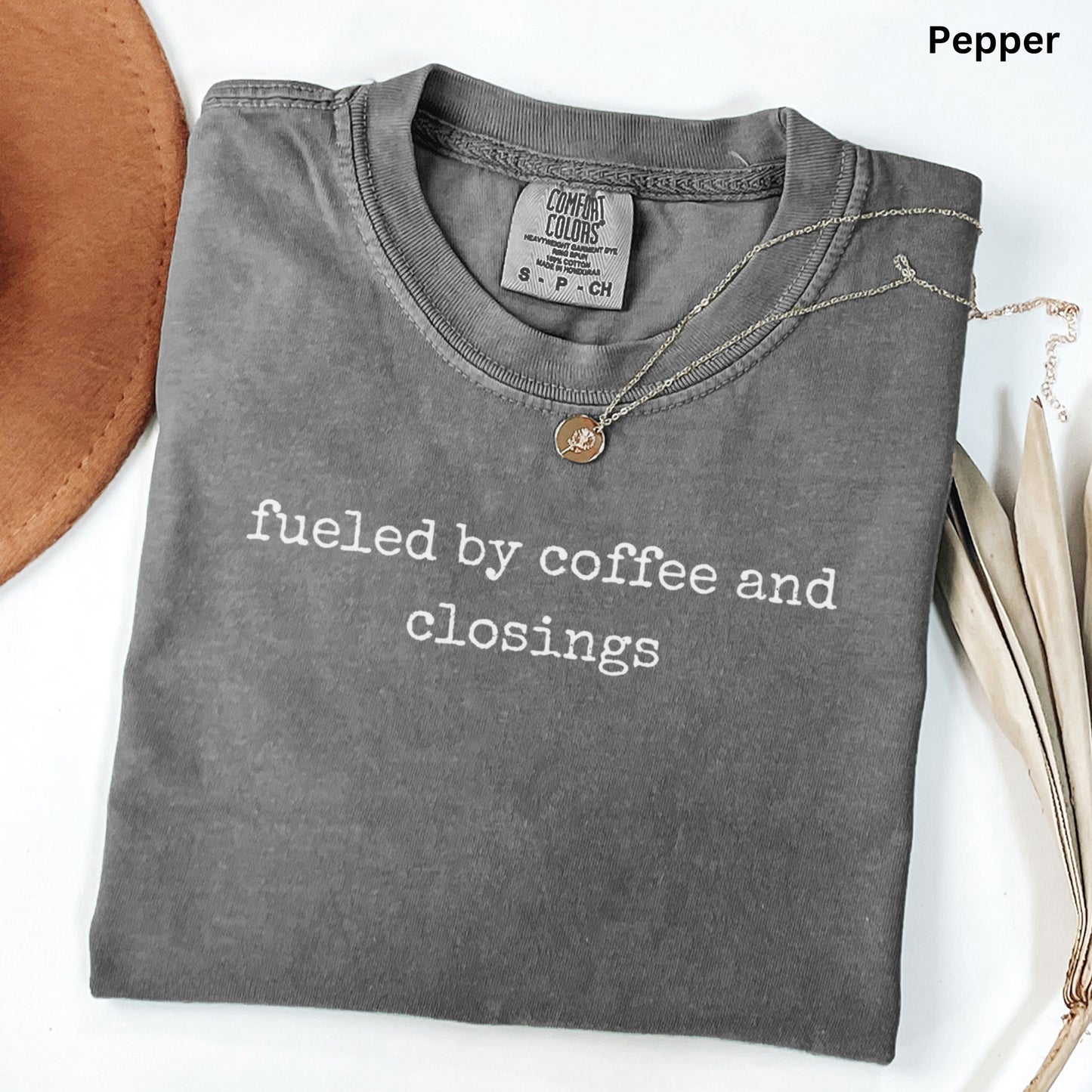 A gray t-shirt with the text "fueled by coffee and closings" printed on it, along with a small pendant necklace.