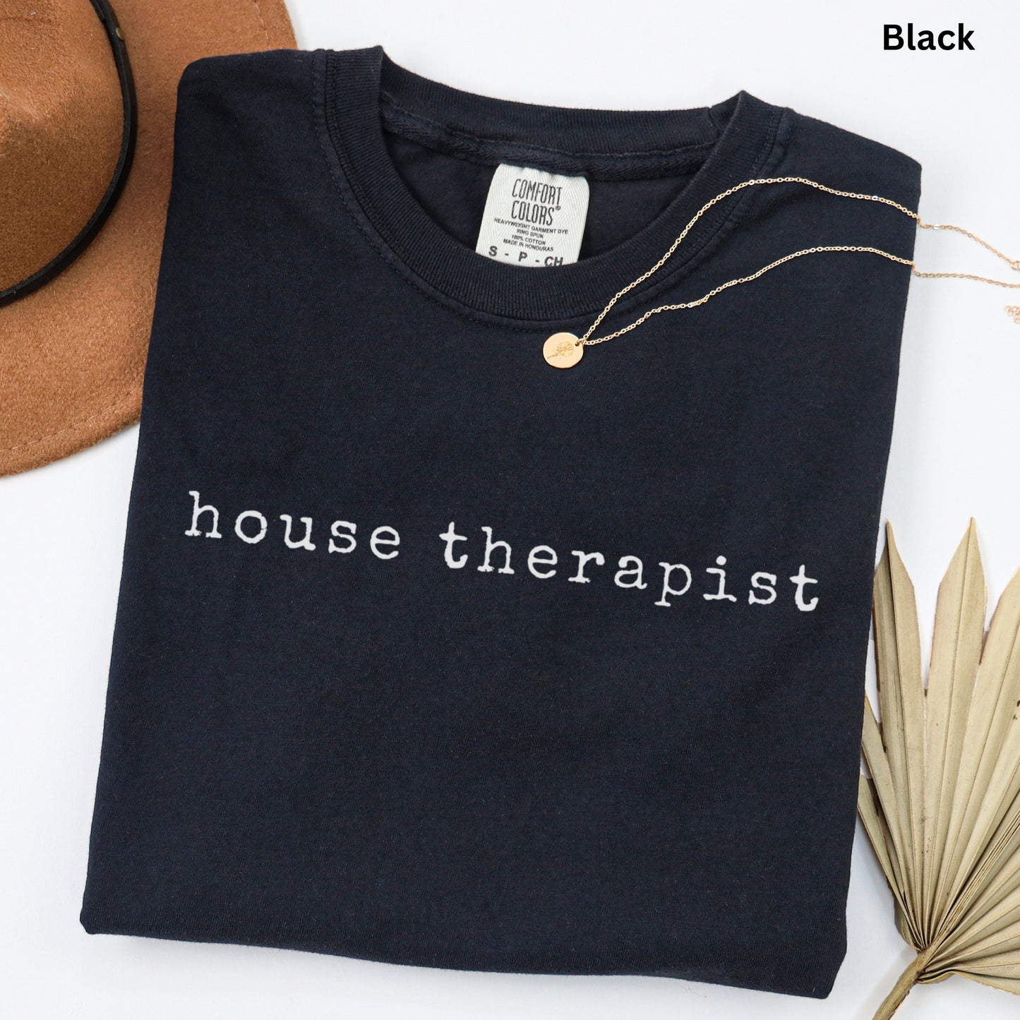 A black t-shirt with the text "house therapist" printed on it, along with a gold necklace and a brown hat.