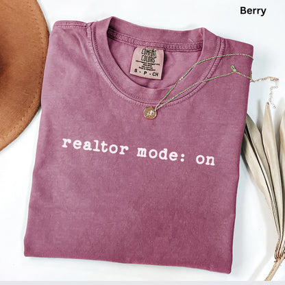 A pink t-shirt with the text "realtor mode: on" printed on it, along with a necklace and a brown hat.