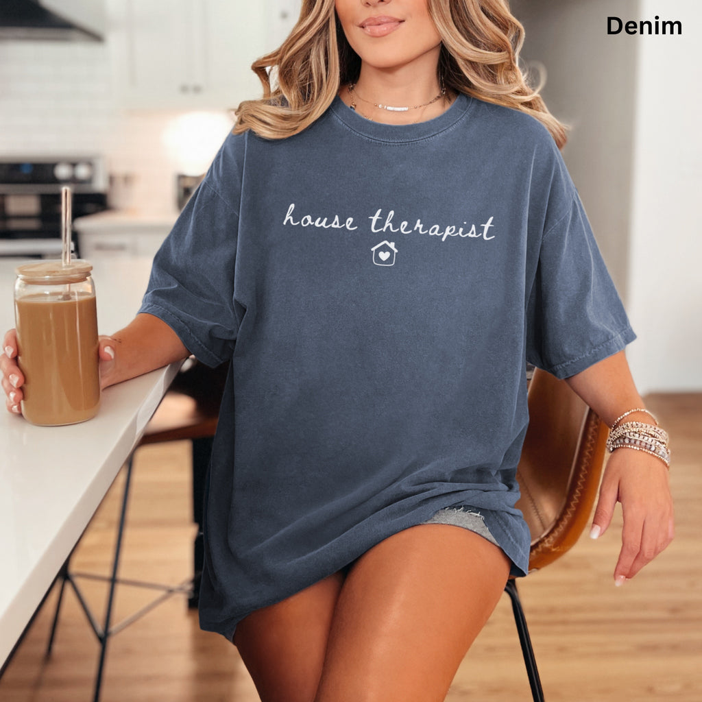 A woman wearing a blue t-shirt with the text "house therapist" is sitting at a kitchen counter, holding a drink in a glass.