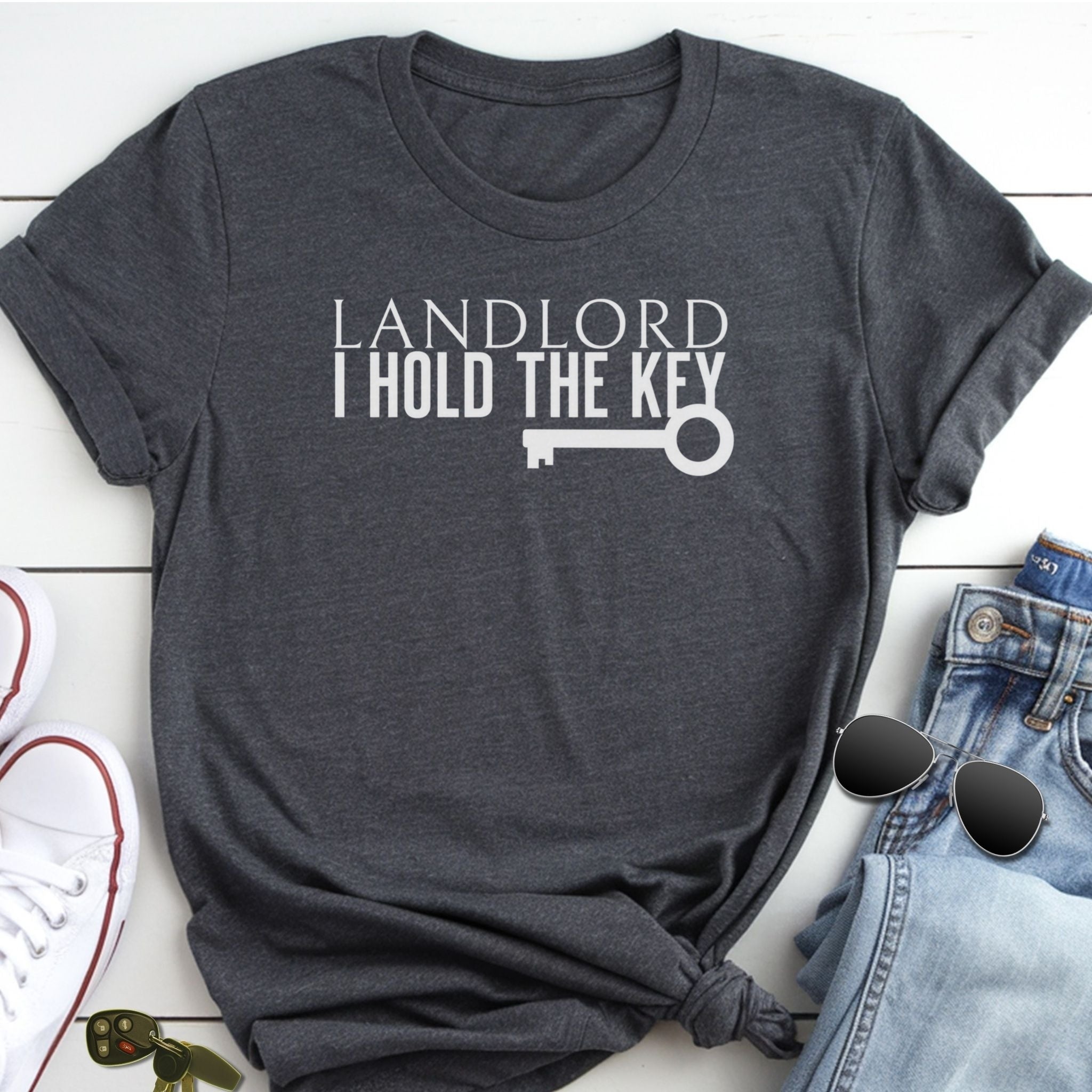 A gray t-shirt with the text "LANDLORD I HOLD THE KEY" printed on it, along with a key graphic. The t-shirt is placed on a white surface, and there are some jeans and sunglasses visible in the background.