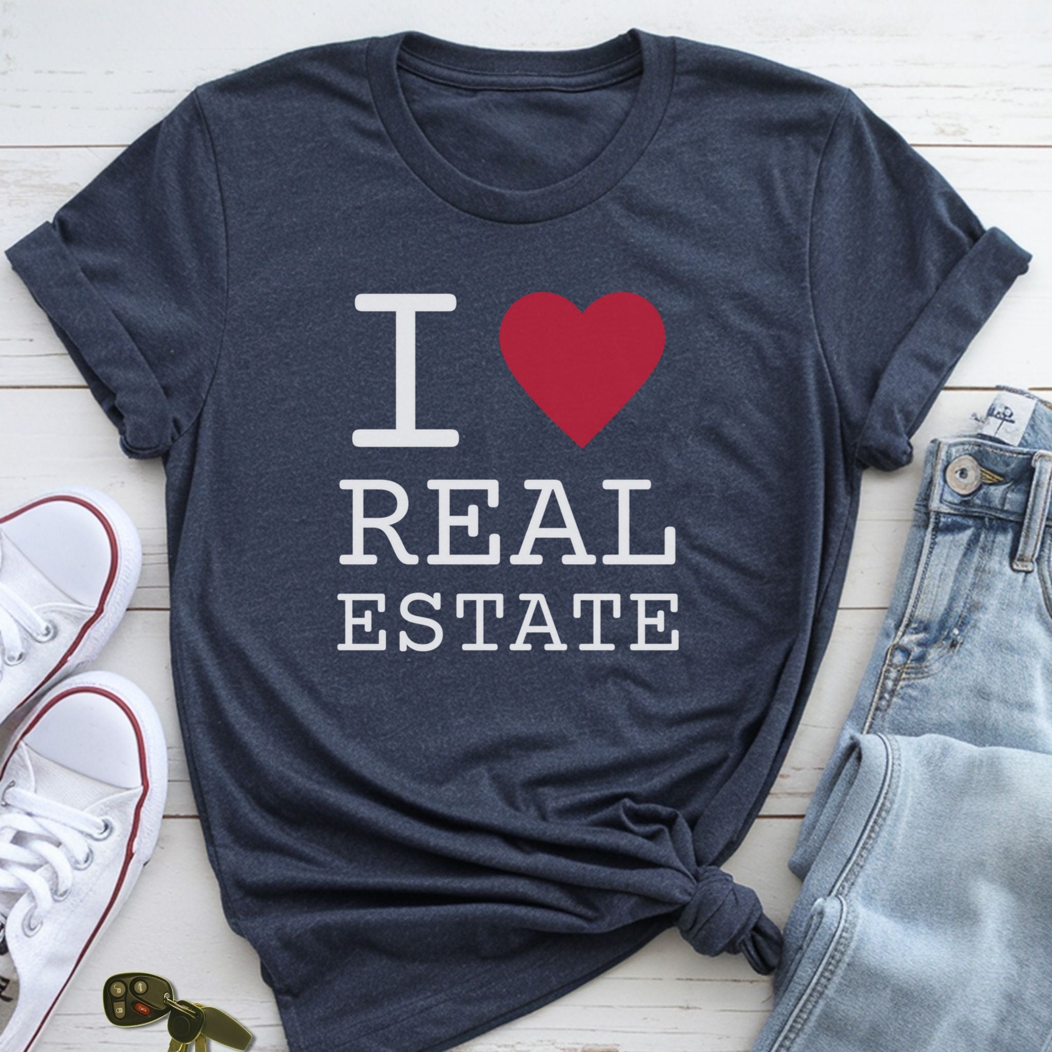 A navy blue t-shirt with the text "I ♥ REAL ESTATE" printed on it, along with a red heart symbol. The t-shirt is laid out on a wooden surface, accompanied by a pair of jeans and a pair of white sneakers.