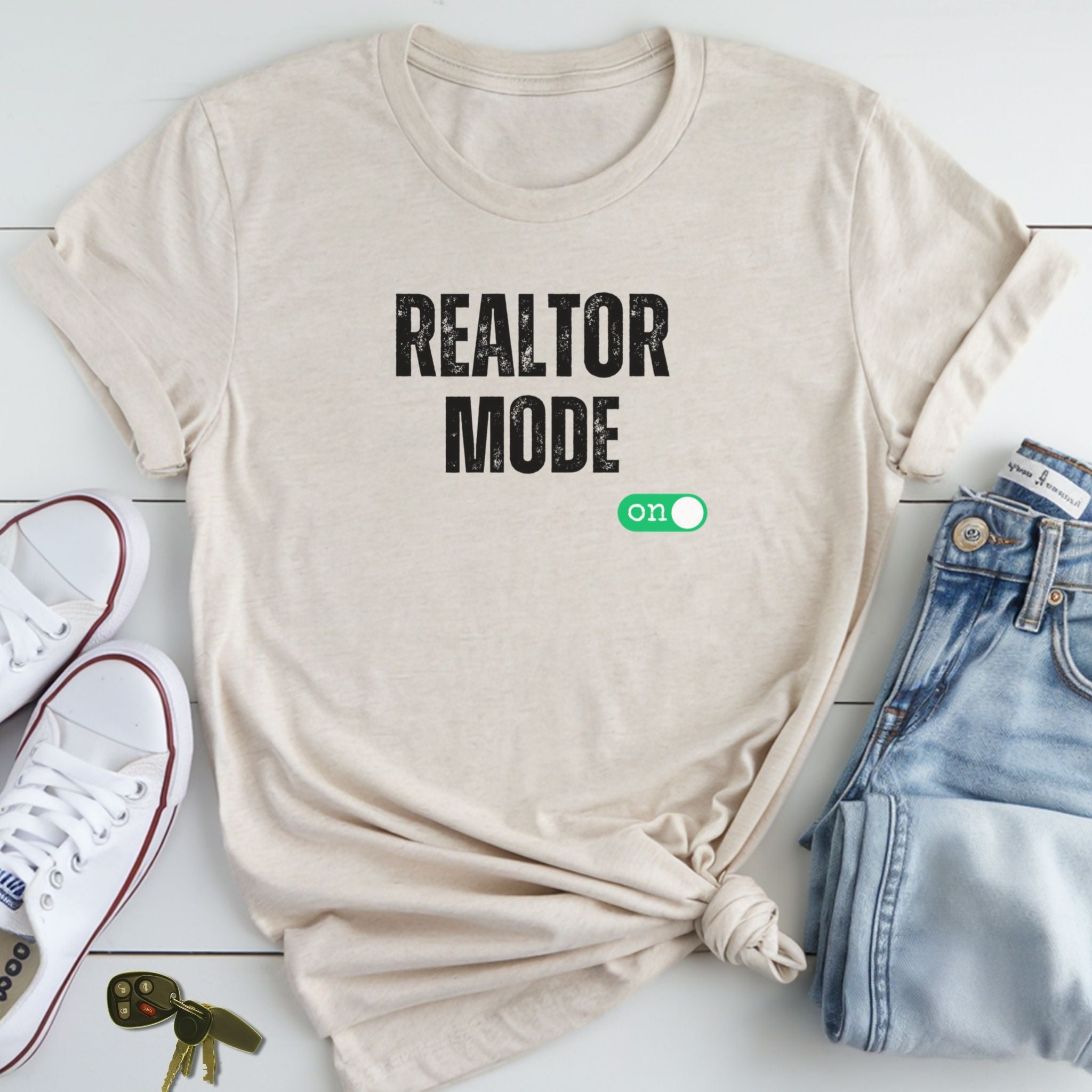 A beige t-shirt with the text "REALTOR MODE" printed on it, along with a green button that says "ON" on a white background.