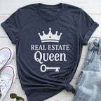 A navy blue t-shirt with the text "REAL ESTATE QUEEN" printed on it, along with a crown and a key, is displayed on a wooden surface alongside a pair of jeans and white sneakers.