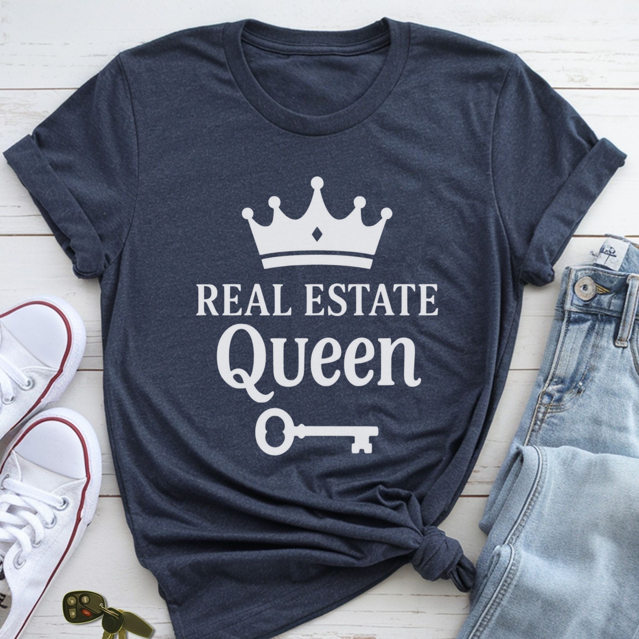 A navy blue t-shirt with the text "REAL ESTATE QUEEN" printed on it, along with a crown and a key, is displayed on a wooden surface alongside a pair of jeans and white sneakers.