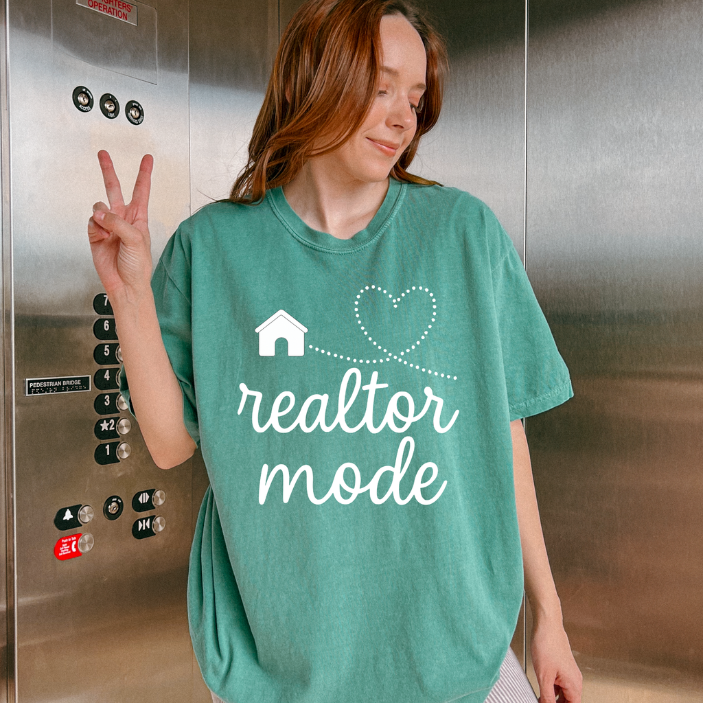 A woman wearing a green t-shirt with the text "realtor mode" and a house graphic is making a peace sign with her right hand.