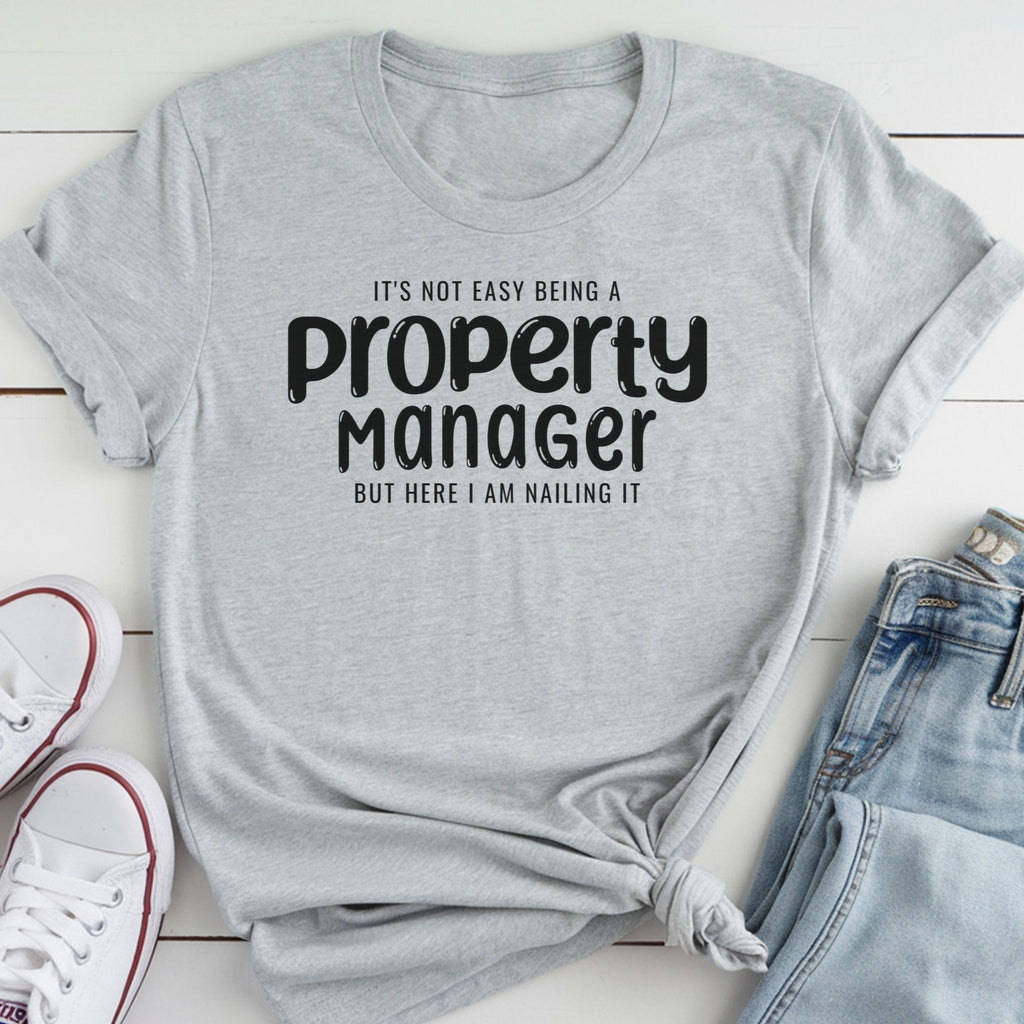 A gray t-shirt with the text "It's not easy being a property manager, but here I am nailing it" printed on it, along with a pair of jeans and white sneakers.