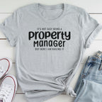 A gray t-shirt with the text "It's not easy being a property manager, but here I am nailing it" printed on it, along with a pair of jeans and white sneakers.