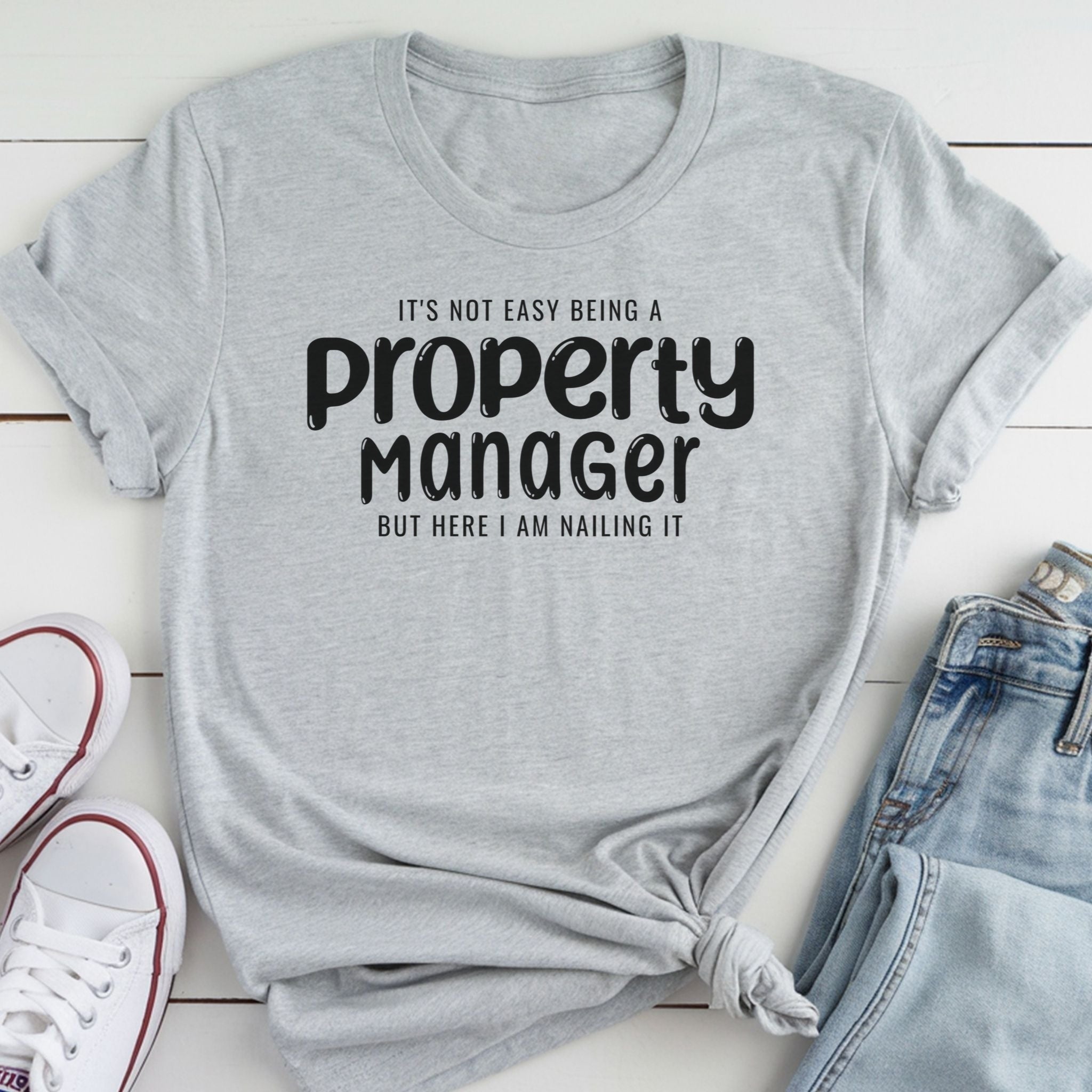 A gray t-shirt with the text "It's not easy being a property manager, but here I am nailing it" printed on it, along with a pair of jeans and white sneakers.