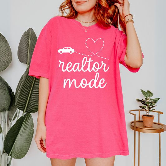 A woman wearing a pink t-shirt with the text "realtor mode" printed on it, standing in front of a plant and a small table.