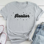 A gray t-shirt with the word "Realtor" printed on it, along with small star-shaped designs, is laid out on a white surface next to a pair of jeans and white sneakers.
