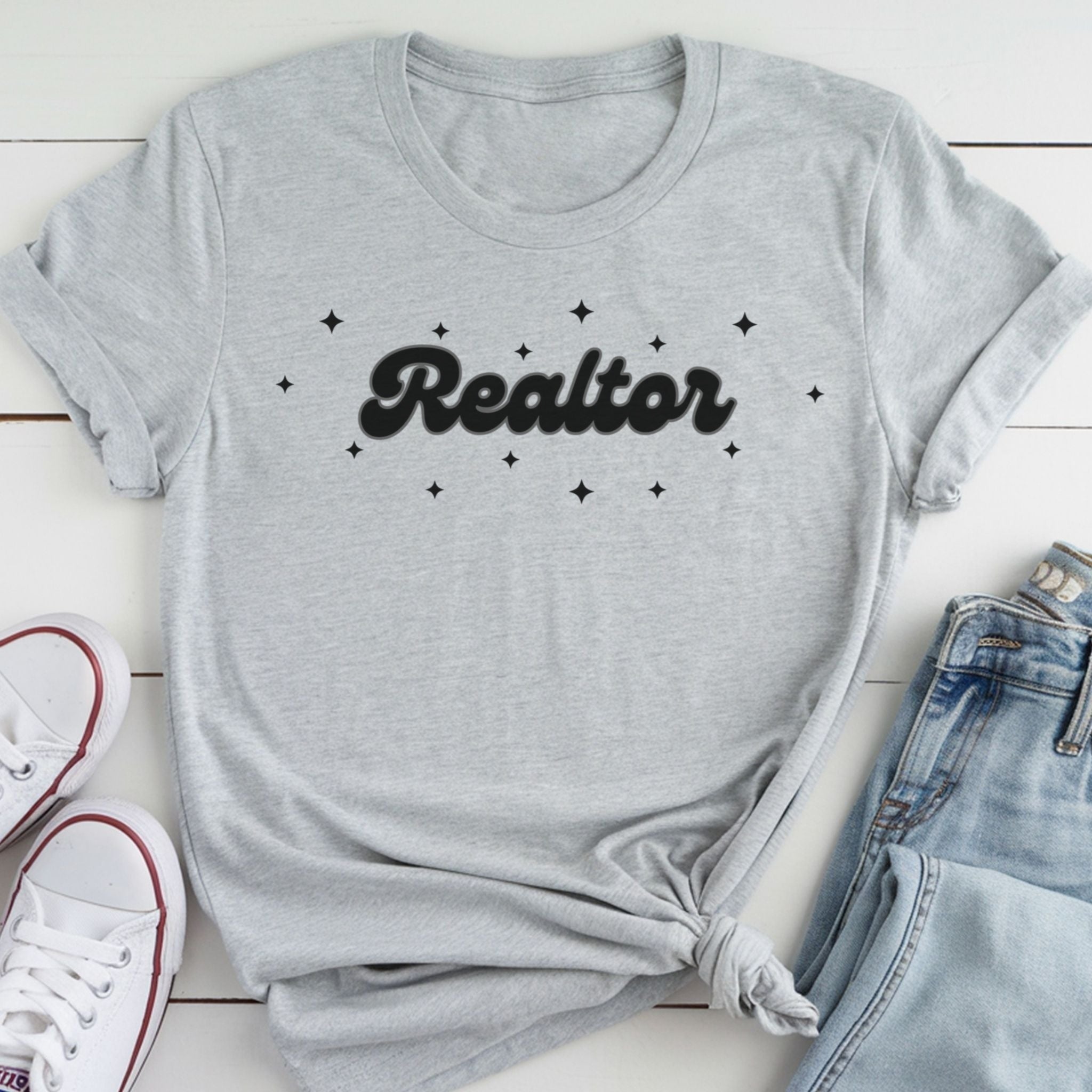 A gray t-shirt with the word "Realtor" printed on it, along with small star-shaped designs, is laid out on a white surface next to a pair of jeans and white sneakers.