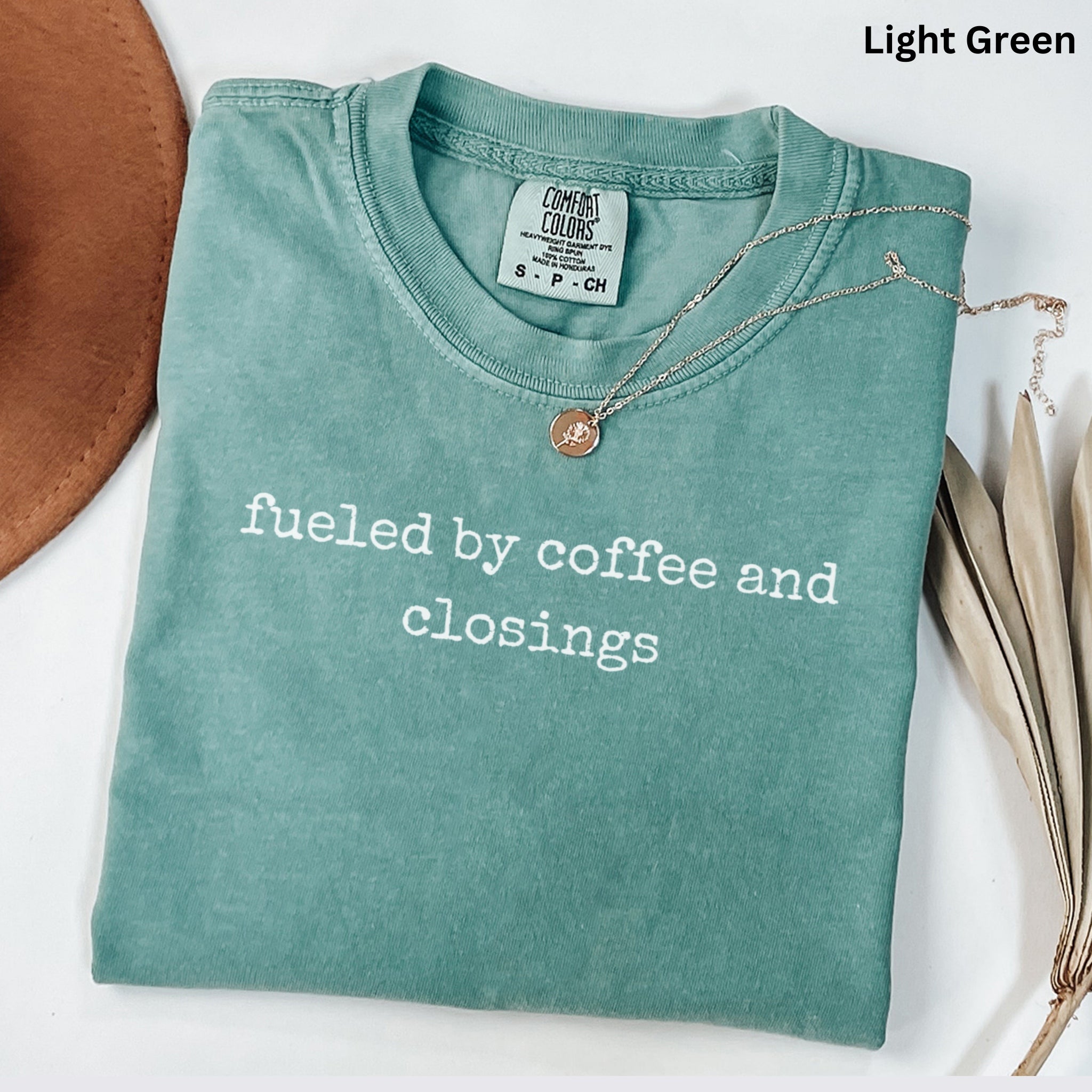 A light green t-shirt with the text "fused by coffee and closings" printed on it, along with a small gold pendant. The t-shirt is laid out on a white surface, and there is a brown hat and some dried leaves nearby.