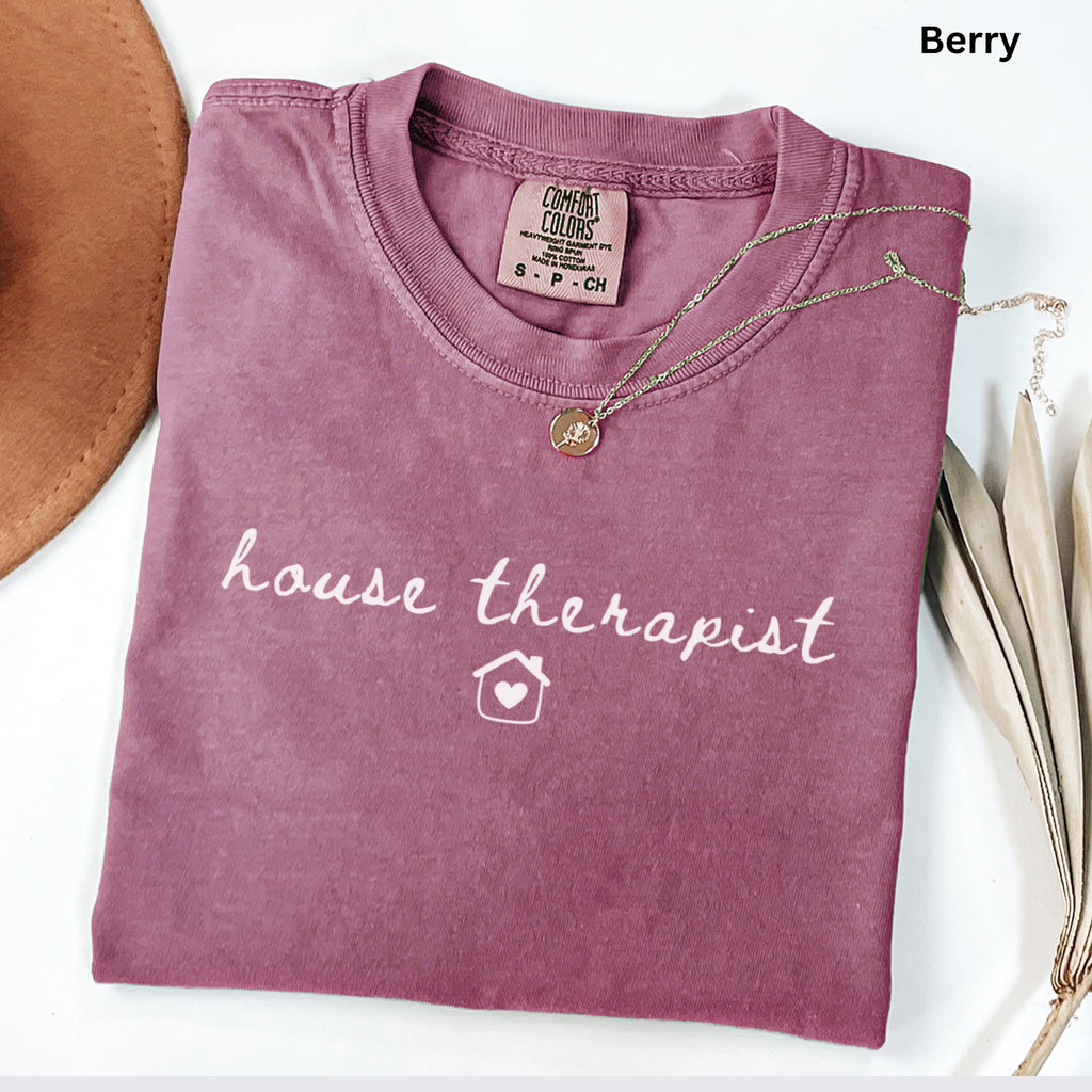 A pink t-shirt with the text "house therapist" printed on it, along with a small house icon. The t-shirt is folded and placed on a white surface, accompanied by a necklace and some leaves.