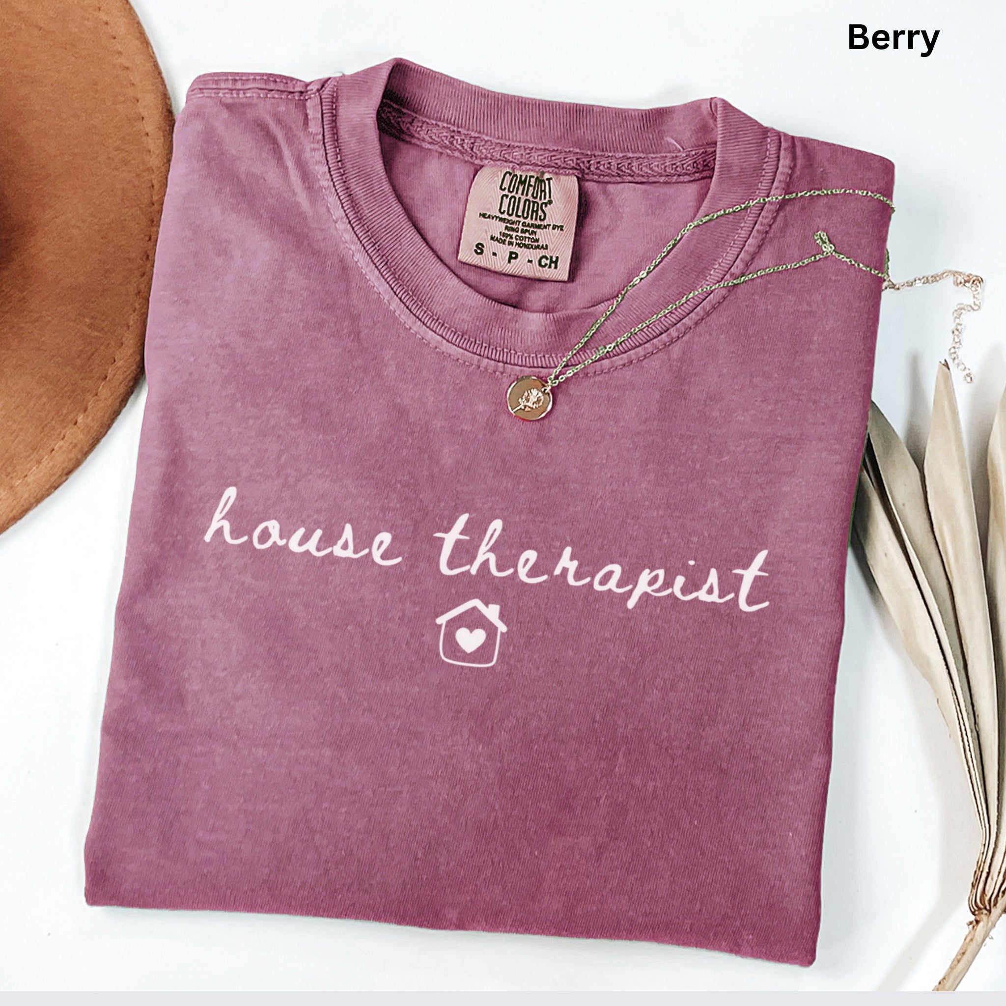 A pink t-shirt with the text "house therapist" printed on it, along with a small house icon. The t-shirt is folded and placed on a white surface, accompanied by a necklace and some leaves.