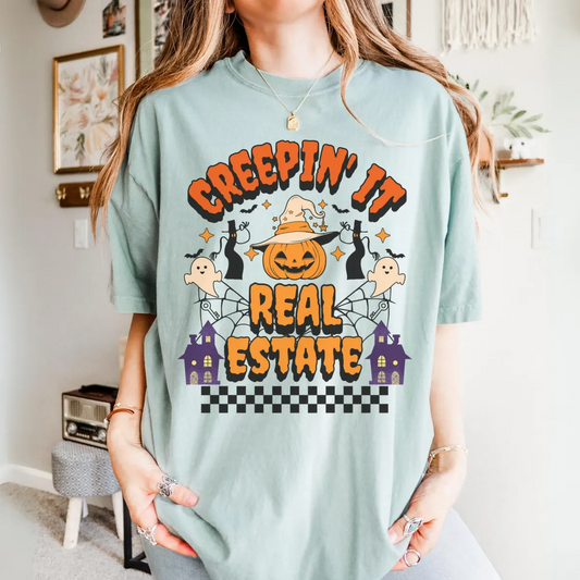 A woman wearing a light blue t-shirt with a Halloween-themed graphic design, including a jack-o-lantern, ghost, spider, and a house.
