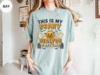 A person wearing a light blue t-shirt with a Halloween-themed design, including a jack-o-lantern, ghost, and houses.