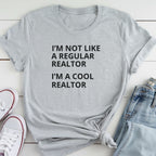 A gray t-shirt with the text "I'M NOT LIKE A REGULAR REALTOR I'M A COOL REALTOR" printed on it, along with a pair of jeans and white sneakers.