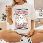 A woman wearing a white t-shirt with a graphic design featuring a ghost and the text "SPOOKY REALATOR" is sitting on a chair in a kitchen.