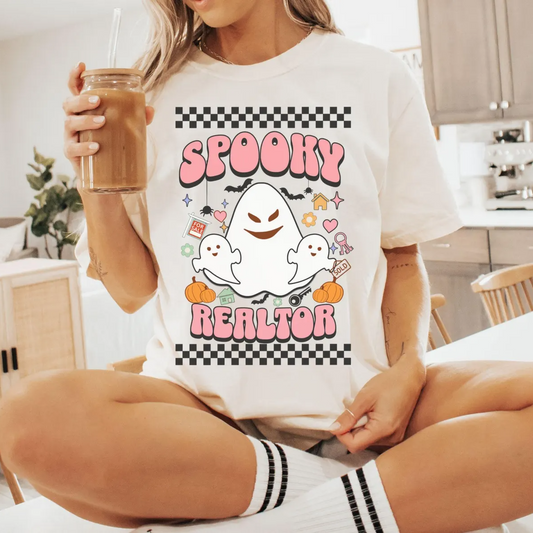 A woman wearing a white t-shirt with a graphic design featuring a ghost and the text "SPOOKY REALATOR" is sitting on a chair in a kitchen.