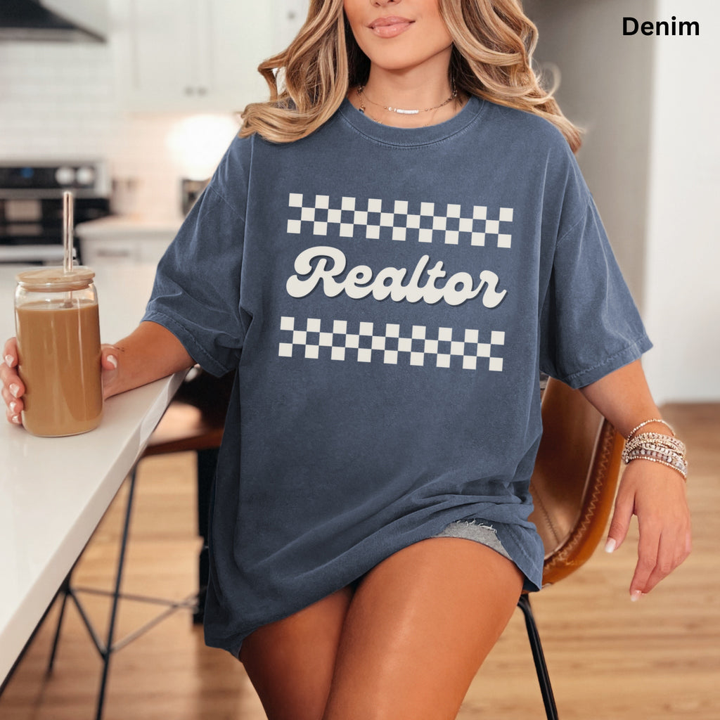 A woman wearing a blue t-shirt with the word "Realtor" printed on it, sitting at a kitchen counter with a drink in a glass.