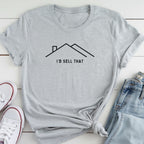A gray t-shirt with a graphic design of a house and the text "I'D SELL THAT" printed on it, along with a pair of jeans and white sneakers.