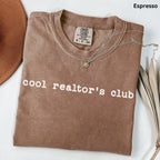 A brown t-shirt with the text "cool realtor's club" printed on it, along with a necklace and some leaves.