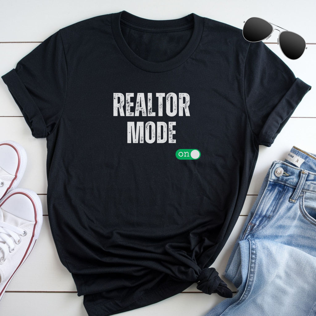 A black t-shirt with the text "REALTOR MODE" printed on it, along with a green button-like symbol. The t-shirt is placed on a white surface, with a pair of sunglasses and a pair of jeans visible in the background.