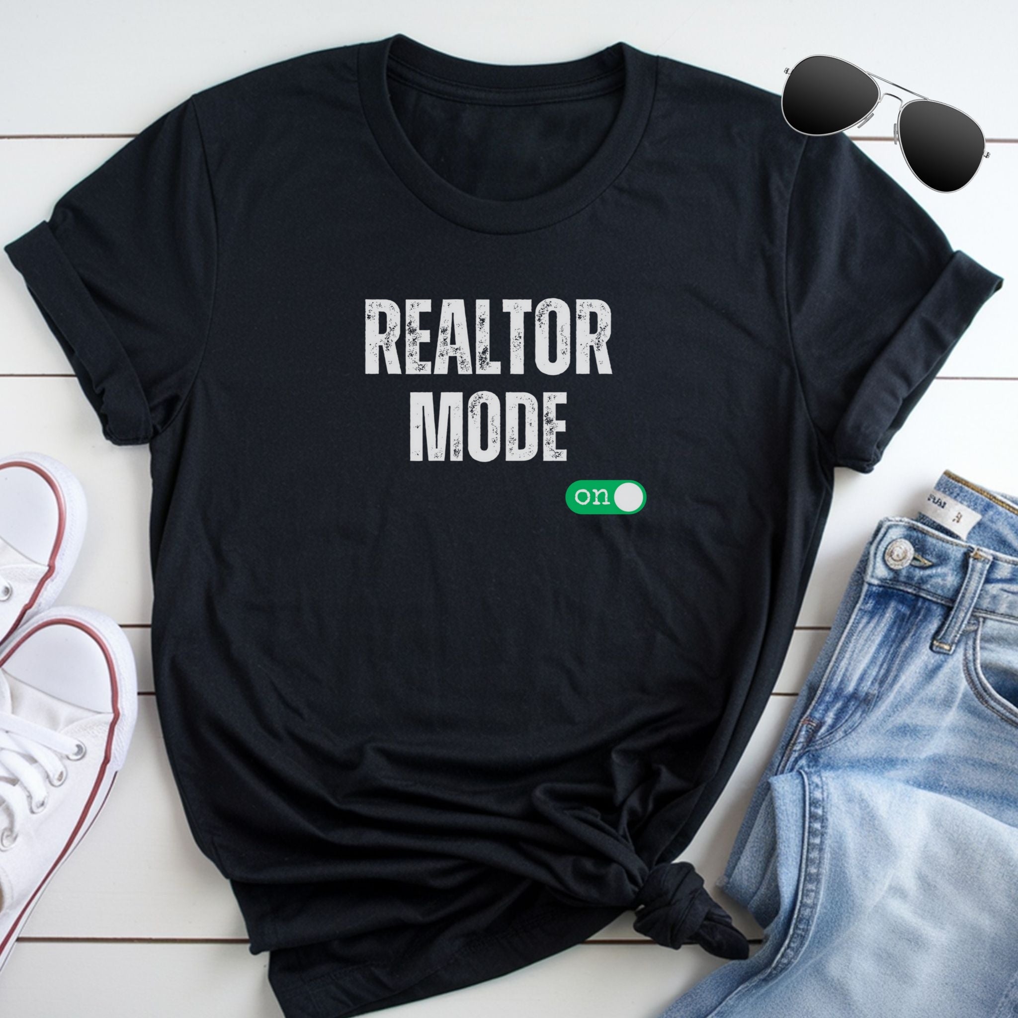 A black t-shirt with the text "REALTOR MODE" printed on it, along with a green button-like symbol. The t-shirt is placed on a white surface, with a pair of sunglasses and a pair of jeans visible in the background.