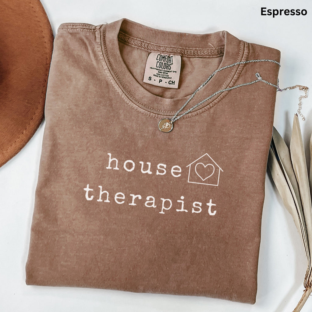 a brown t-shirt with the text "house therapist" printed on it, along with a heart symbol and a house icon. The t-shirt is folded and placed on a white surface, with a brown hat and some leaves visible in the background.