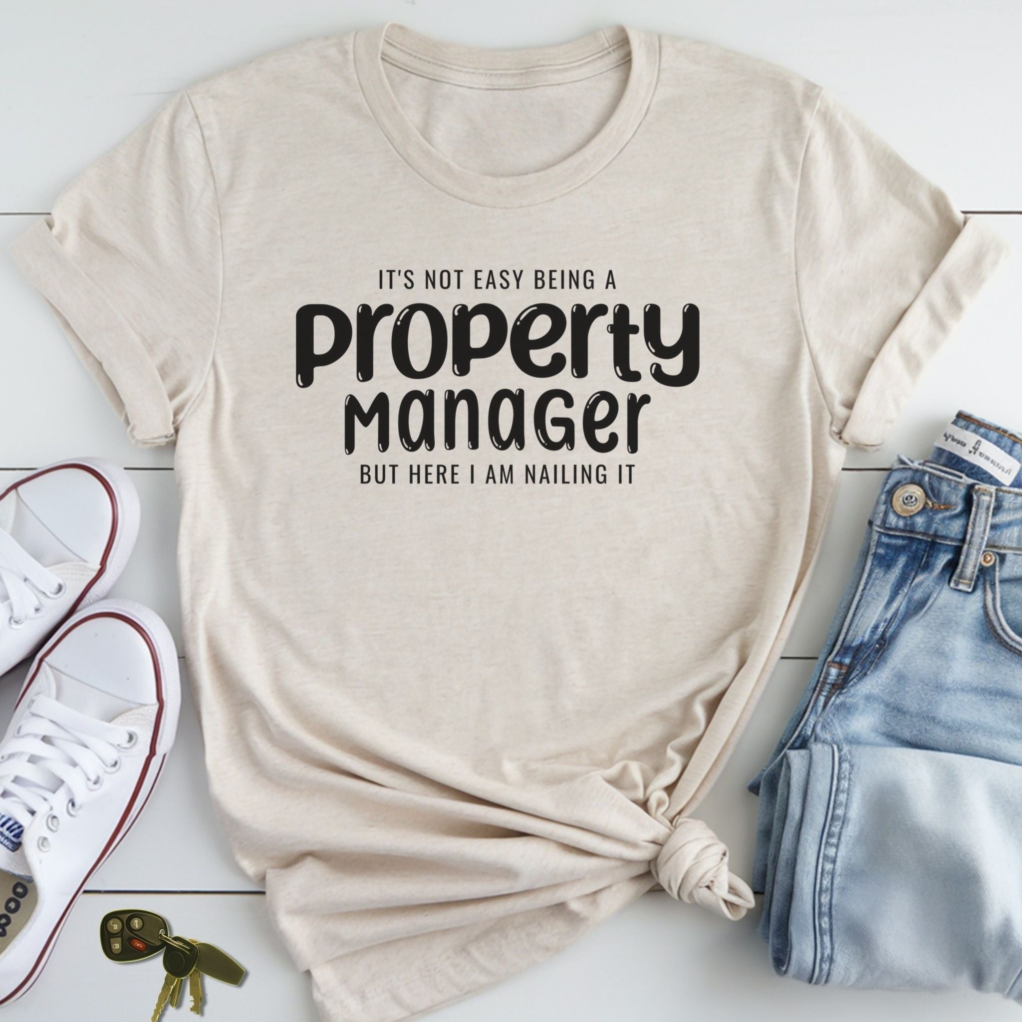 A beige t-shirt with a humorous text printed on it, along with a pair of jeans and sneakers, all laid out on a white surface.