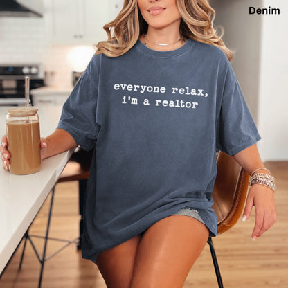 A woman wearing a blue t-shirt with the text "Everyone relax, I'm a realtor" is sitting at a kitchen counter, holding a drink.