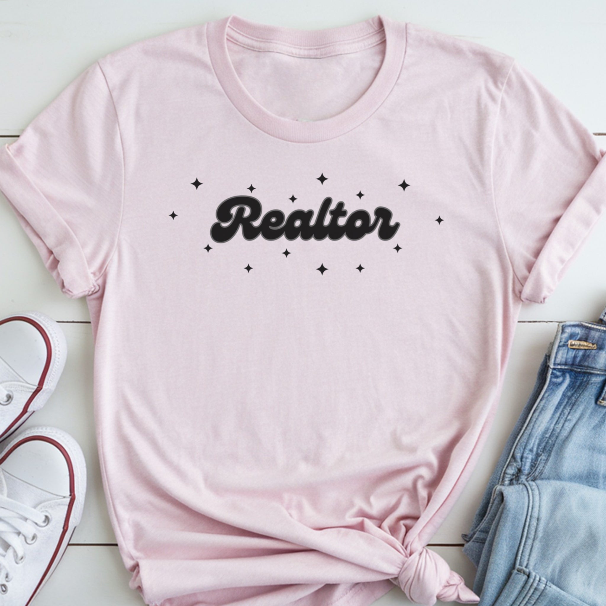 A pink t-shirt with the word "Realtor" printed on it, along with small star-shaped designs, is laid out on a white surface next to a pair of jeans and white sneakers.