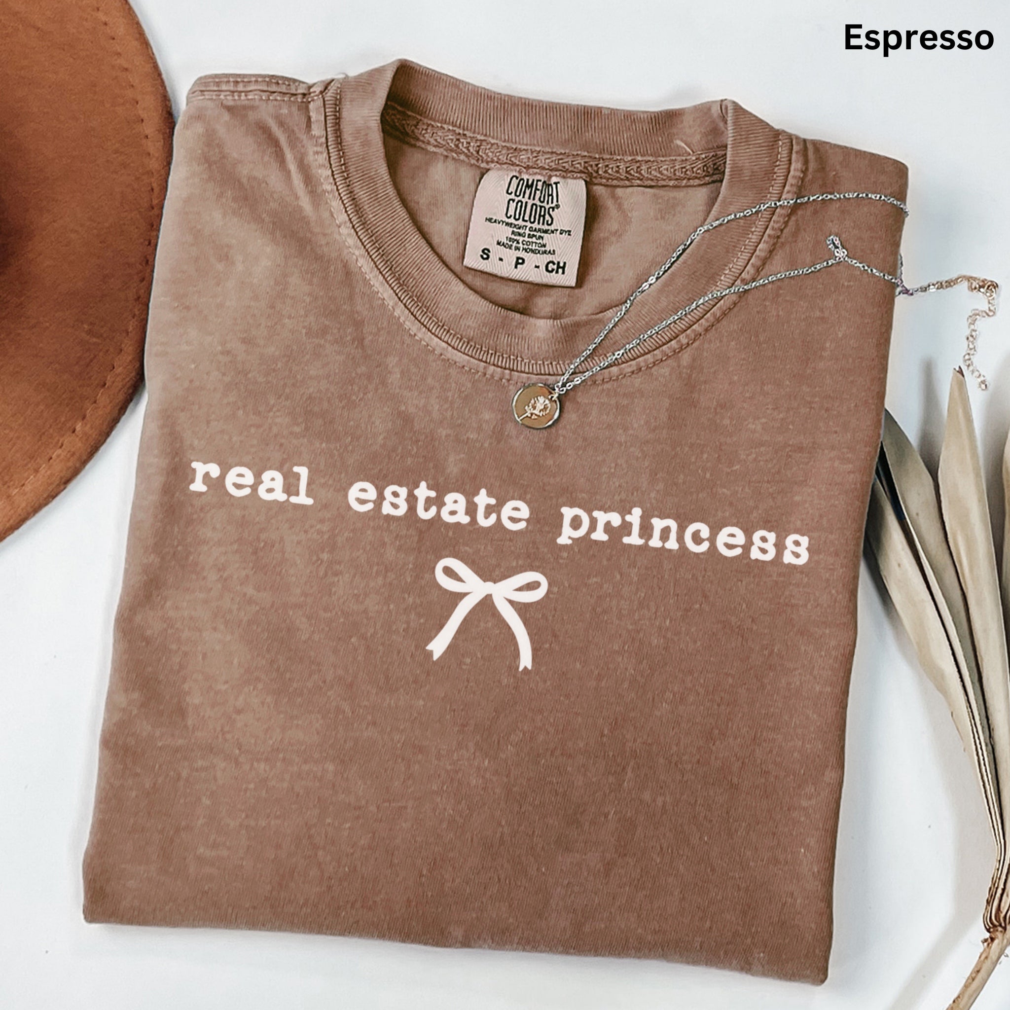a brown t-shirt with the text "real estate princess" printed on it, along with a ribbon design. The t-shirt is laid out on a white surface, and there is a necklace and some leaves nearby.