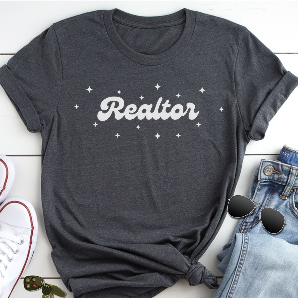 A gray t-shirt with the word "Realtor" printed on it, along with small white stars, is displayed on a white surface alongside a pair of jeans and sunglasses.