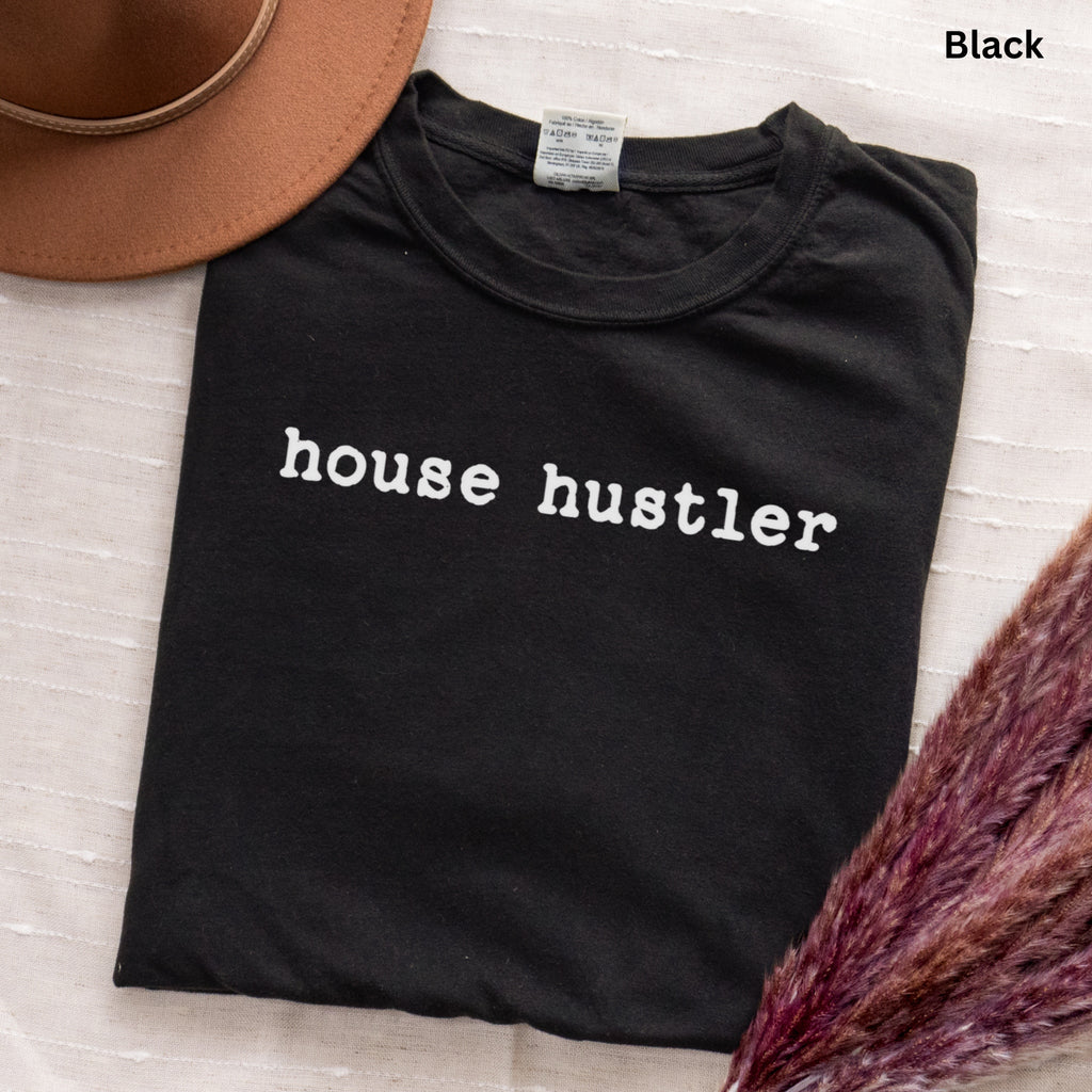 A black t-shirt with the text "house hustler" printed on it, along with a brown hat and some purple flowers.