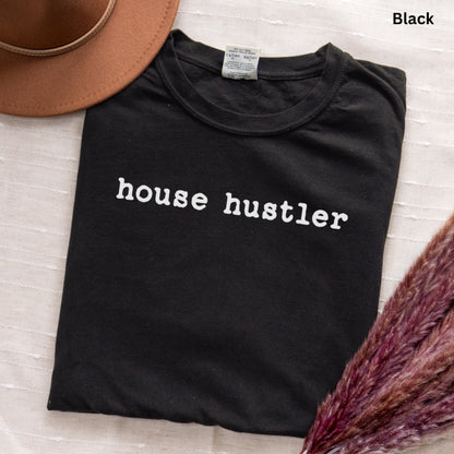 A black t-shirt with the text "house hustler" printed on it, along with a brown hat and some purple flowers.