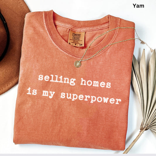 a red t-shirt with the text "selling homes is my superpower" printed on it, along with a gold necklace and a brown hat.
