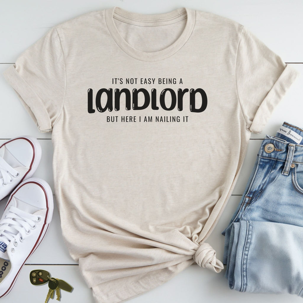 A beige t-shirt with the text "It's not easy being a landlord but here I am nailing it" printed on it, along with a pair of jeans and a pair of white sneakers.