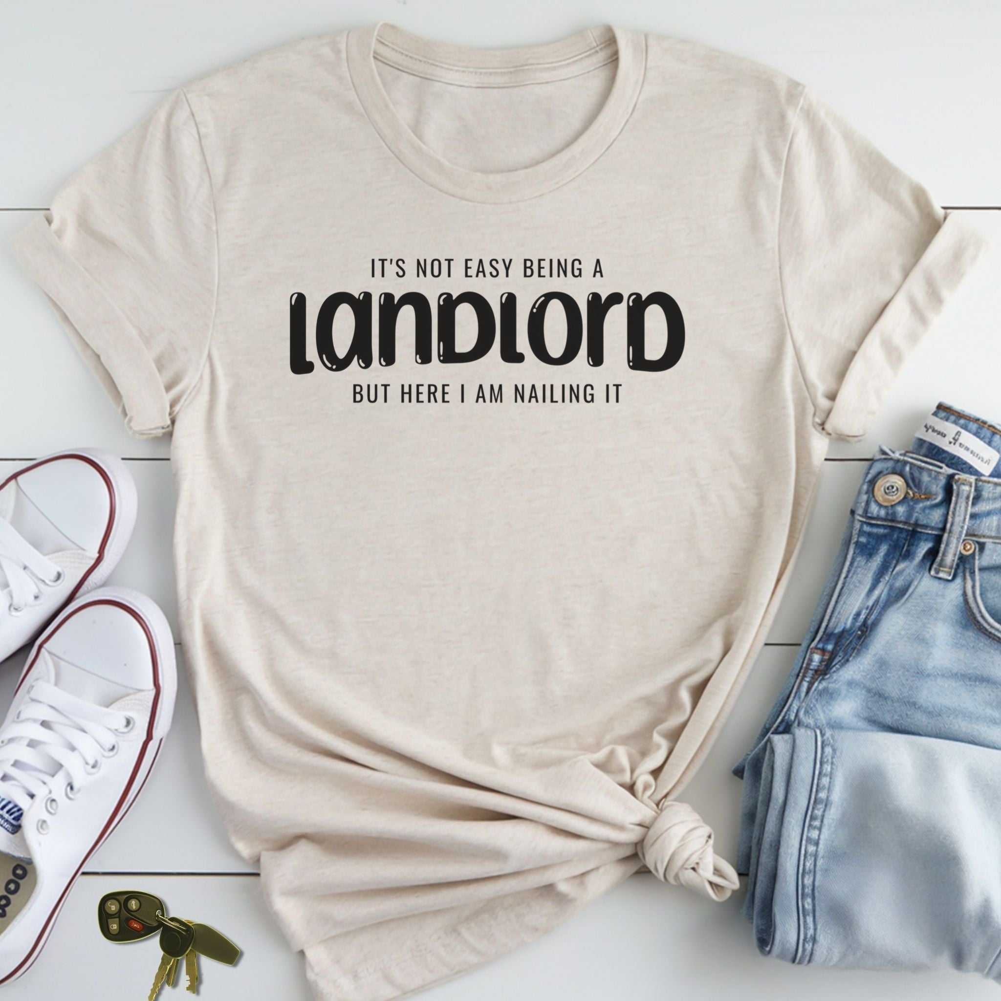 A beige t-shirt with the text "It's not easy being a landlord but here I am nailing it" printed on it, along with a pair of jeans and a pair of white sneakers.